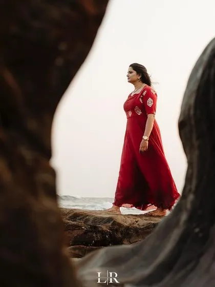 A creative shot of a woman in a red dress walking along the rocks by the sea, framed by natural elements.