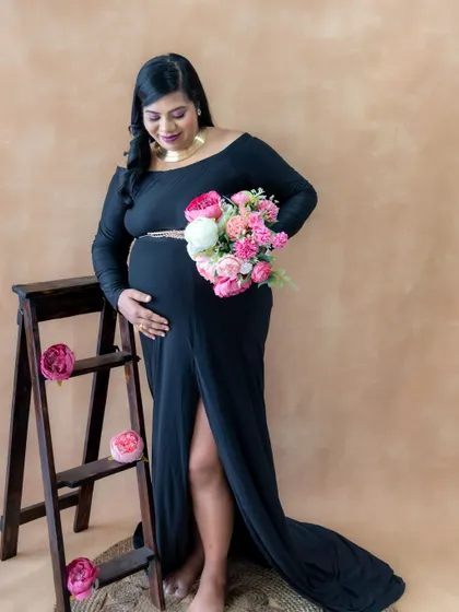 A classic studio portrait with a simple, warm-toned backdrop. The mom-to-be looks elegant in a black gown, holding a bouquet of flowers.