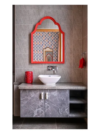 A pop of color in this bathroom, with a bright red arched mirror against a grey marble-finish vanity and patterned wallpaper reflected in the mirror.