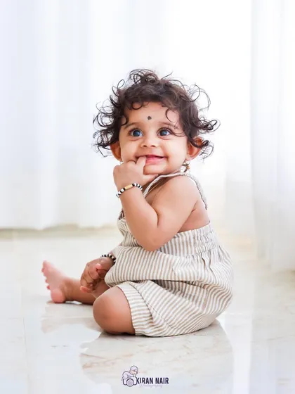 An incredibly sweet portrait of a baby girl with curly hair and bright eyes, sitting on a reflective studio floor. Her charming expression is the centerpiece of this simple and clean shot.