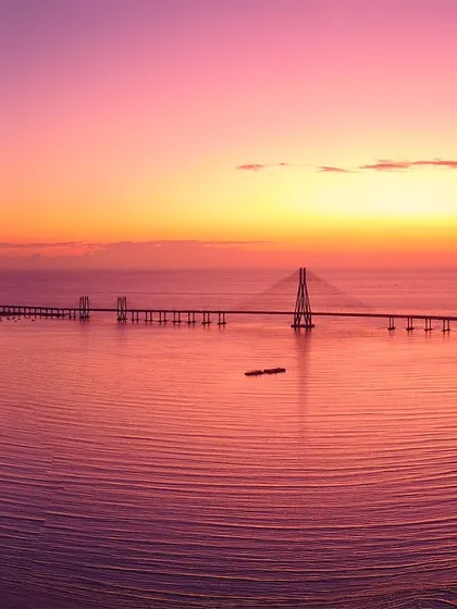 A breathtaking aerial view of the Bandra-Worli Sea Link in Mumbai, silhouetted against a fiery pink and orange sunset reflecting on the calm sea.