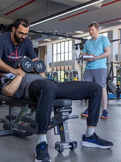 An assessor observes a dumbbell press, evaluating the trainee's control and execution.