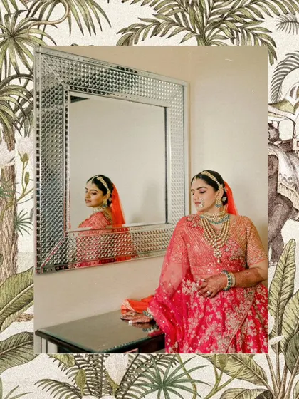 A moment of reflection for the bride. The ornate mirror frame complements her royal bridal look, featuring a heavily embroidered red lehenga and exquisite layered green bead jewelry.