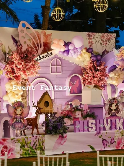 A magical fairy garden party for Nishvika, glowing under the evening sky. This enchanting setup includes a whimsical purple fairy house, large butterfly wing props, custom name lettering, and soft lighting from hanging lanterns to create a dreamlike atmosphere.