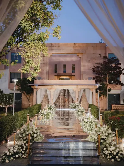 An all-white Varmaala setup featuring a mirrored walkway leading to a rustic wooden mandap. The path is lined with lush arrangements of white chrysanthemums, creating a serene and elegant aisle against the backdrop of the Jodhpur venue.