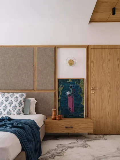 **Cocoon House, Ahmedabad:** In the bedroom, we designed a custom headboard that combines fabric panels and wood trim, integrating the bedside table and artwork into a single, cohesive element. This detail-oriented approach creates a clean, uncluttered, and personalized space.
