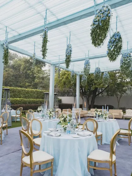 The guest seating area for the Anand Karaj, featuring elegant tables with blue linens and hanging floral installations overhead. The clear-top tent allows for an open, airy feel.