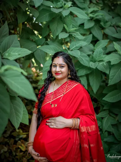 A close-up of the mother-to-be in her vibrant red saree, surrounded by lush green leaves. Her expression is one of serene confidence and joy.
