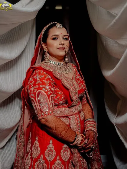 A powerful bridal portrait. The bride stands confidently, her red lehenga vibrant and her gaze direct, framed by flowing white curtains.