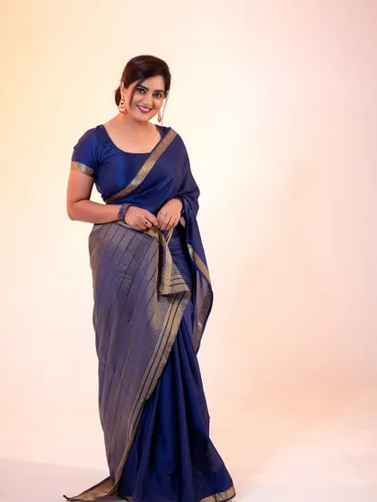 A full-length studio portrait in a classic blue saree. The clean, warm-toned background ensures the subject stands out, highlighting the simple elegance of the attire.