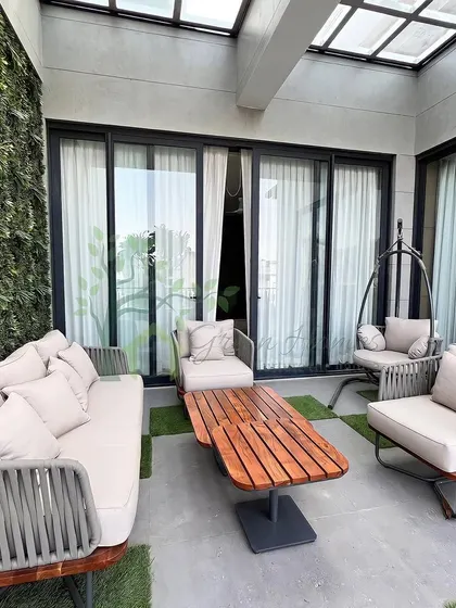 A photograph of a finished balcony project in Yojna Vihar. This enclosed space features a glass skylight, a vertical green wall, and comfortable woven furniture, turning a small balcony into a cozy, private retreat.