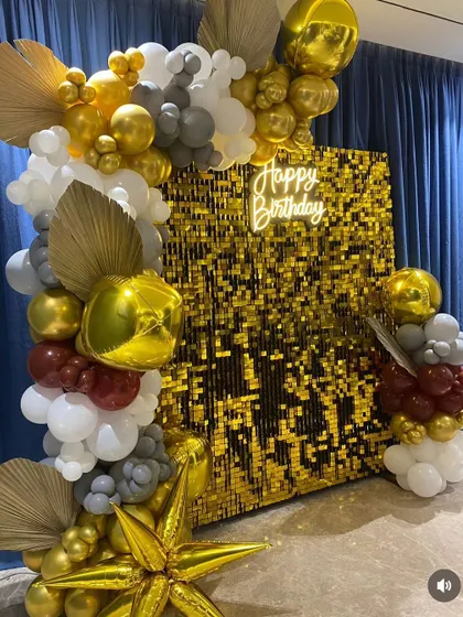 A sophisticated birthday backdrop with a gold shimmer wall and a unique balloon arrangement in white, grey, red, and gold, accented with dried palm leaves.