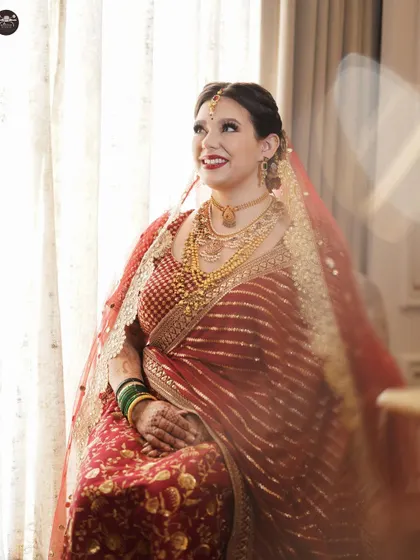 A candid portrait of a bride looking joyfully away from the camera, bathed in soft window light. Her traditional red saree and elaborate gold jewelry shine in this moment of happiness.