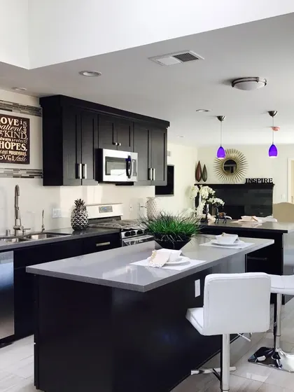 A dark and dramatic open-plan kitchen. The black shaker-style cabinets and grey countertops are paired with a central island and bar stools, creating a social hub within the home.