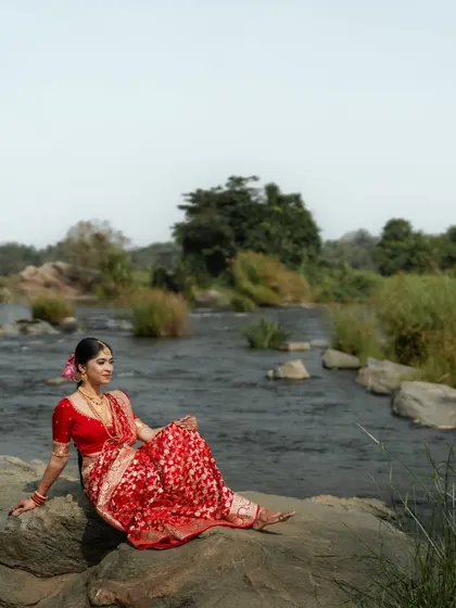 An artistic outdoor shot of a Coorgi bride in a red saree, seated by a river. The natural setting beautifully contrasts with her vibrant traditional attire.