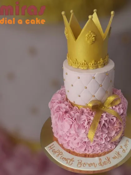 A luxurious two-tier princess cake in pink and gold. The bottom tier has a beautiful ruffled texture, while the top is quilted and finished with a golden crown.