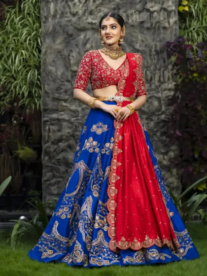The full view of this red and blue raw silk lehenga showcases its grandeur. The gold zardosi work catches the light beautifully, and the contrasting red dupatta adds another layer of richness to this bridal outfit.
