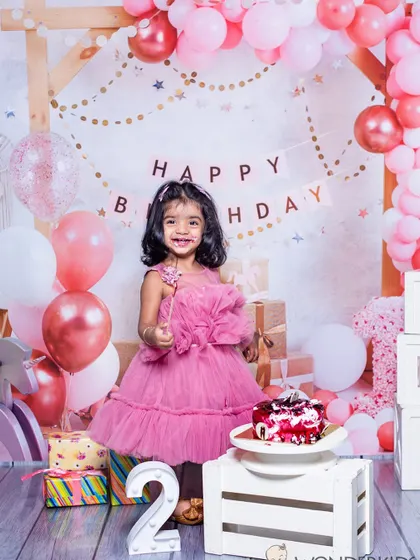A little girl celebrating her second birthday with a big smile. The pink balloon arch, presents, and birthday cake create a perfect party scene for her special day.