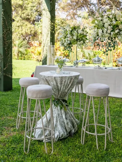 A stylish high-top table with modern bar stools, set on the lawn for guests to gather. The patterned tablecloth adds a touch of subtle elegance.