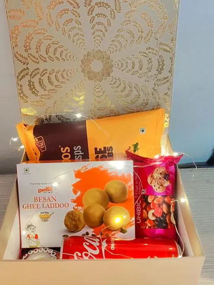 A corporate hamper that balances traditional and modern tastes. It includes classic besan ghee laddoos, savory snacks, a can of Coca-Cola, and a decorative diya.