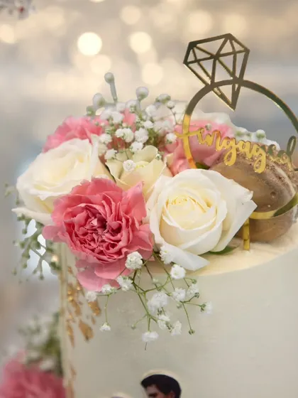 A close-up of the floral topper on the engagement cake, featuring fresh roses, carnations, and an elegant "Engaged" ring topper.