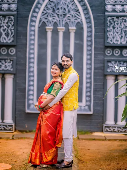 A full-length portrait of the expecting couple in their traditional outfits. The intricate wall behind them adds a touch of architectural elegance to this beautiful and culturally rich maternity photograph.
