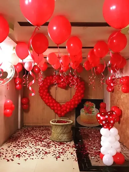 A complete room transformation designed for a big romantic gesture. The ceiling is covered with red balloons, a large heart made of smaller balloons serves as the centerpiece, and the floor is blanketed in red rose petals.