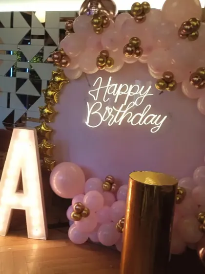 I can personalize your birthday decorations with light-up marquee letters for initials, as shown here with the letter 'A'. This setup is paired with a pink and gold balloon arch and a 'Happy Birthday' neon sign.