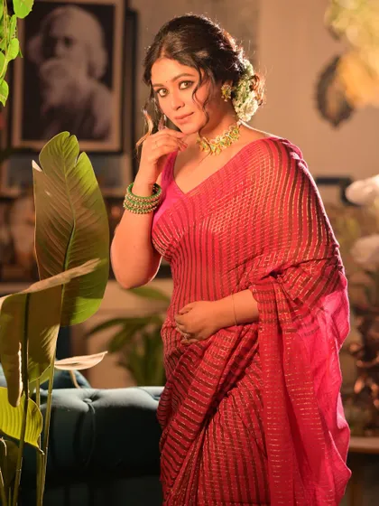 A festive portrait for 'Shubho Noboborsho' (Bengali New Year). The model is dressed in a shimmering pink saree, with her hair adorned with flowers, set in a warmly lit, decorated room.
