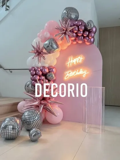 A trendy and fun birthday setup with a pink arched backdrop and neon sign. The balloon arrangement includes pink, white, and metallic balloons, accented with disco balls and starburst foils for a party vibe.