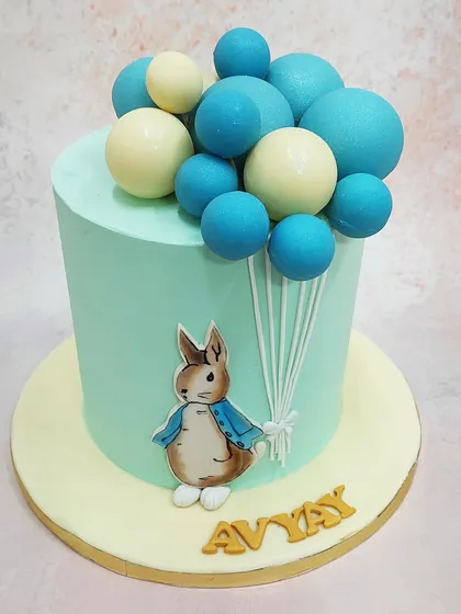 We bring a classic tale to the party with this charming Peter Rabbit cake. The beloved character is featured on a soft, mint-green tier, holding a large bouquet of blue and white balloons, creating a whimsical and gentle design for a first birthday or baby shower.