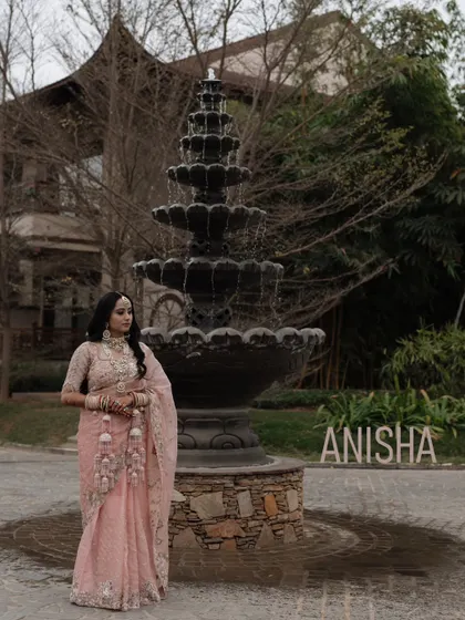 A full-length portrait of the bride in a delicate pink saree, posing elegantly in front of a grand fountain.
