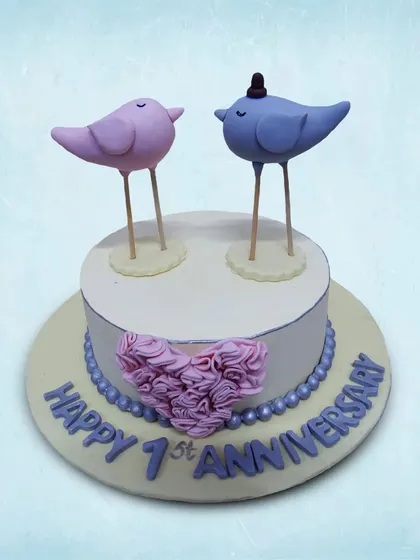 We celebrate a first anniversary with these adorable lovebirds. Two sweet bird figures, one pink and one blue, stand atop a simple cake adorned with a heart of roses, symbolizing a year of love and partnership.