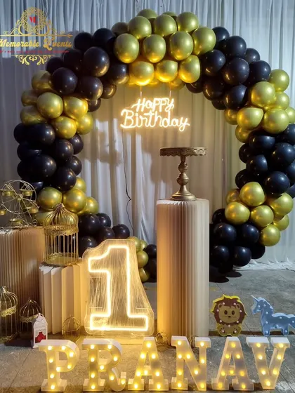 A classic black and gold first birthday setup with a full balloon ring, light-up number '1', and marquee letters for the name.