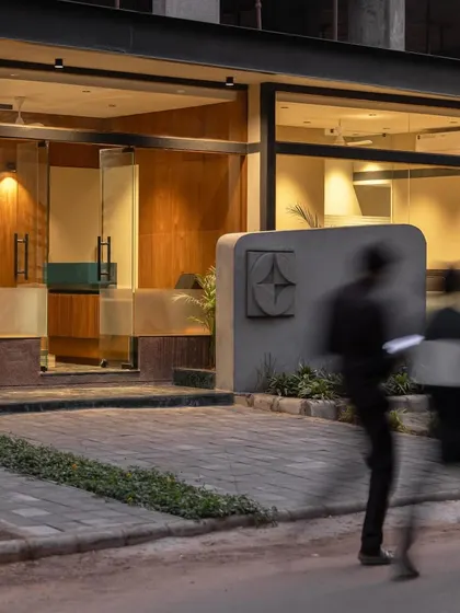 **Sanctum Site Office, Ahmedabad:** An evening shot captures the warm, inviting glow from within the office. The use of motion blur with people walking by emphasizes that this is a space of activity, a prelude to the community that will soon inhabit the residences.