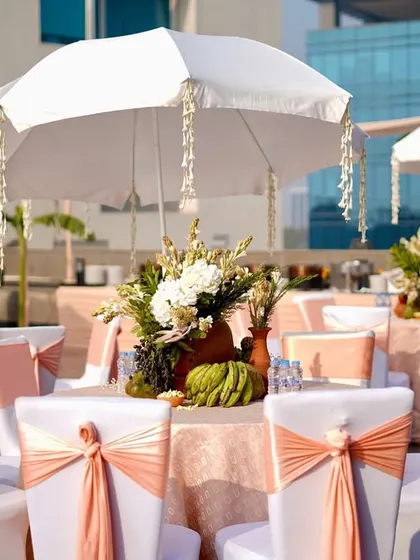 An elegant outdoor dining area with tables dressed in white and soft peach. The umbrellas, draped with hanging jasmine garlands, add an ethereal and traditional touch.