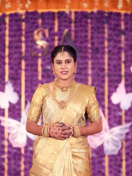 A full-length portrait of the bride in a stunning gold saree, standing against a vibrant purple and butterfly-themed stage.