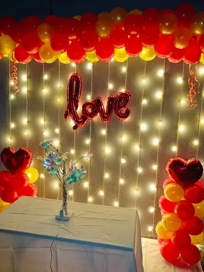 A romantic terrace setup with a fairy light wall. The decor includes a 'Love' foil balloon and red and yellow balloon columns.
