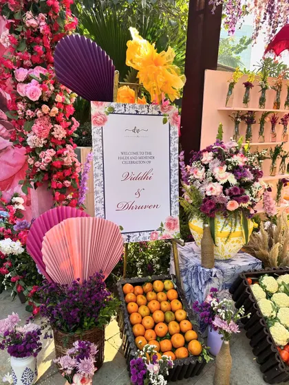 The welcome area for a Haldi and Mehendi celebration, overflowing with colorful flowers, fresh oranges, and a personalized sign for the couple.