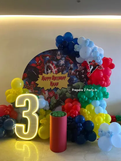 An action-packed Avengers theme for a third birthday party. This setup includes a circular backdrop with all the superheroes, a light-up marquee number, and a balloon garland in the Avengers' signature colors.