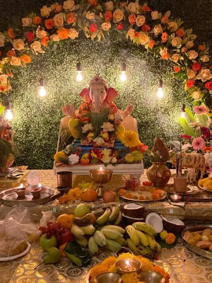 A rustic and earthy Ganpati decoration with a green foliage backdrop and an arch of orange and white flowers. The exposed Edison bulbs add a warm, vintage touch to the festive setup.