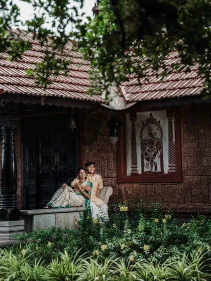 A serene portrait of a couple sitting on the steps of a traditional house, framed by lush greenery and beautiful architecture.