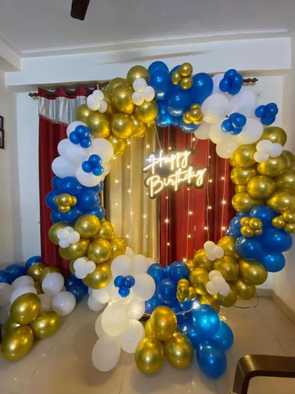 A birthday balloon ring in blue, white, and gold, with a "Happy Birthday" neon sign.
