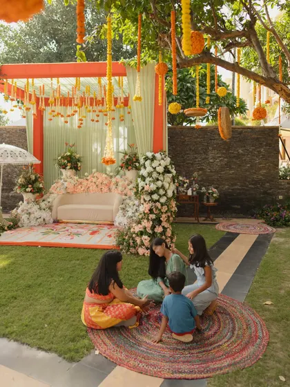 A relaxed moment at a daytime Mehendi, showing the guest seating area with colorful jute rugs and a stage decorated with pastel florals and mint green drapes.