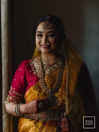A classic bridal portrait by a window. The soft, natural light enhances her features and the rich colors of her yellow and pink silk saree.