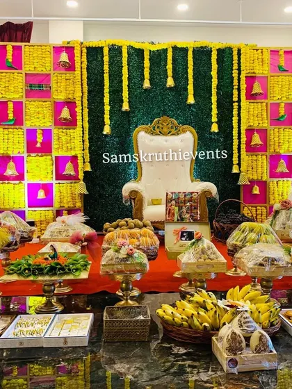 A bright and festive Seemantha backdrop with a checkered pattern of yellow flowers and bells against a green background. The offering plates are neatly wrapped, ready for the ceremony.