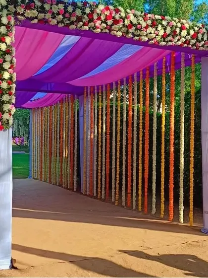A grand outdoor wedding entrance tunnel created with purple and pink drapes and lined with hanging strings of colorful flowers.