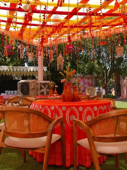 A view of the guest tables, featuring bright red patterned tablecloths and cane-back chairs. The centerpiece of rustic terracotta vases adds an earthy, traditional touch to the vibrant Haldi decor.