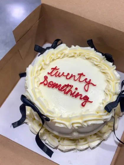 This "twenty something" bento cake is a perfect example of the trendy vintage buttercream style. It features elegant off-white piping and delicate black ribbon bows for a chic and celebratory look.