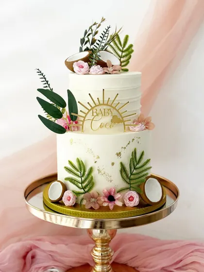A trendy, boho-chic baby shower cake with a "Baby Coco" theme. This two-tiered cake is decorated with fondant coconuts, tropical leaves, and delicate flowers.